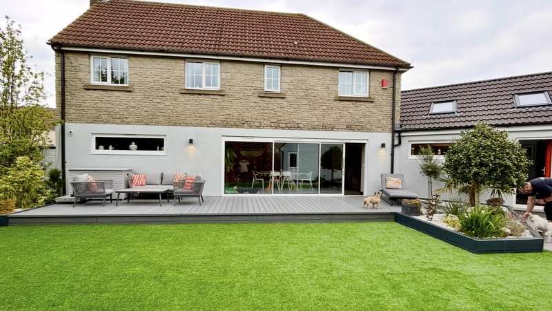The same house after a single-storey rear extension in white render, with a tiled roof, rooflights, decking and a planted border
